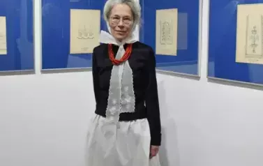 Kobieta w stroju ludowym pozująca na tle rysunk&oacute;w ze wzorami haft&oacute;